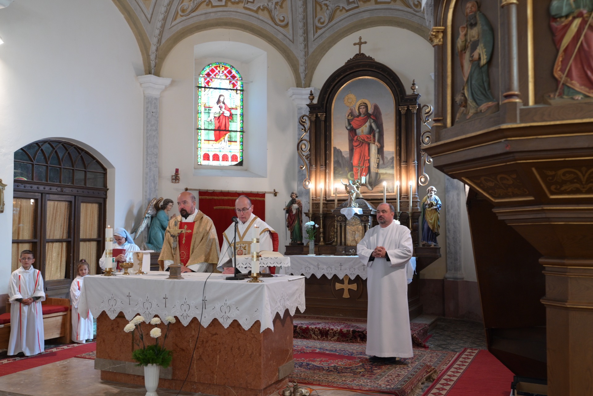 Katolikus templomban misén álló papok, oltárterítővel, színes üvegablakkal és aranyozott szoborral a háttérben.